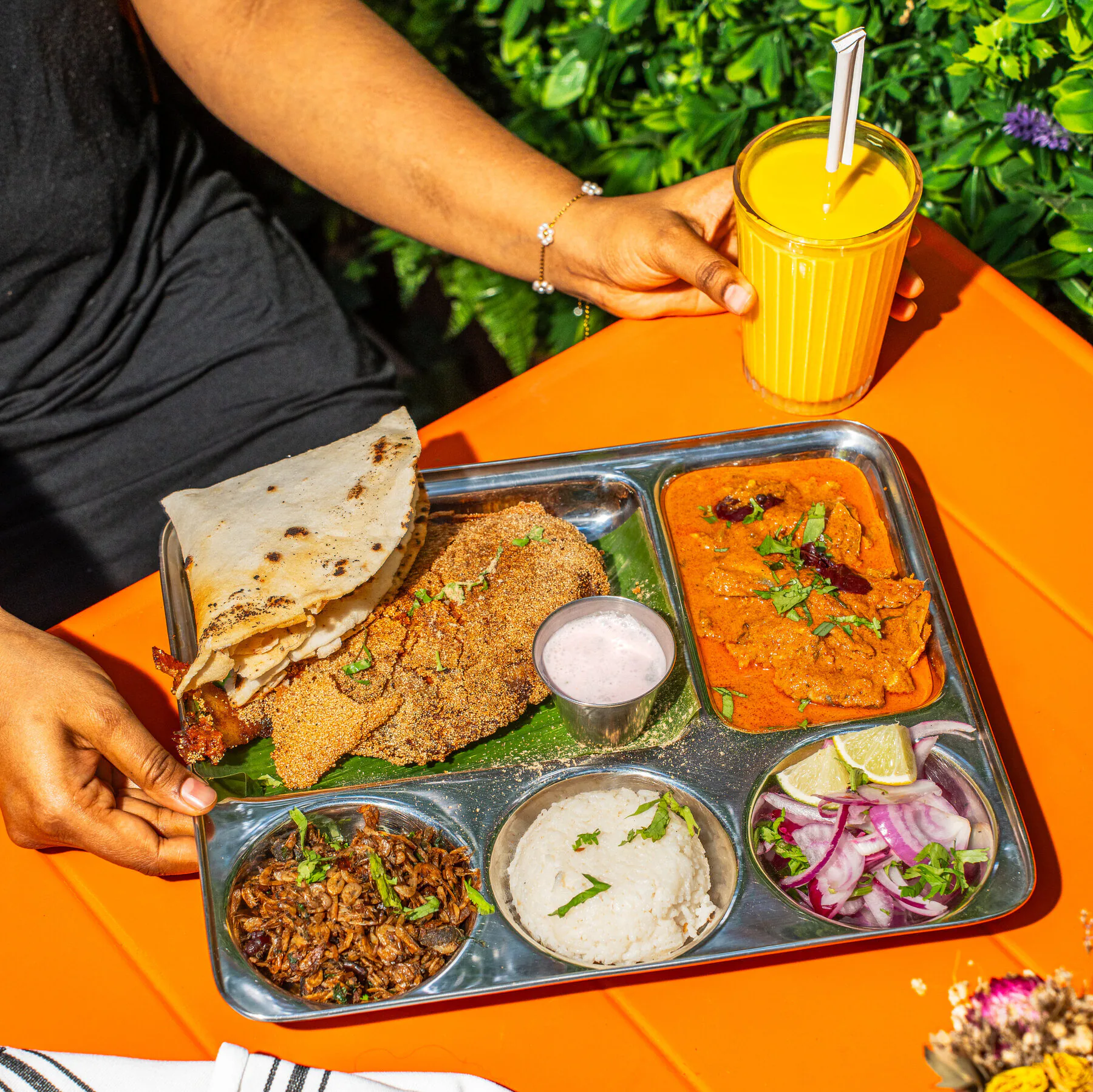 A plated Indian meal with flatbread, curry, sides, and a mango drink set out on a bright table for a shower-style gathering