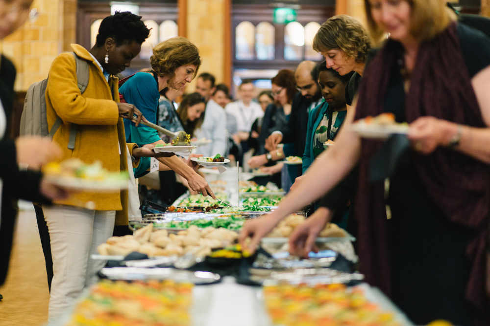 Guests serving themselves from a community-style buffet table set with salads, buns, and hot items during a campus event