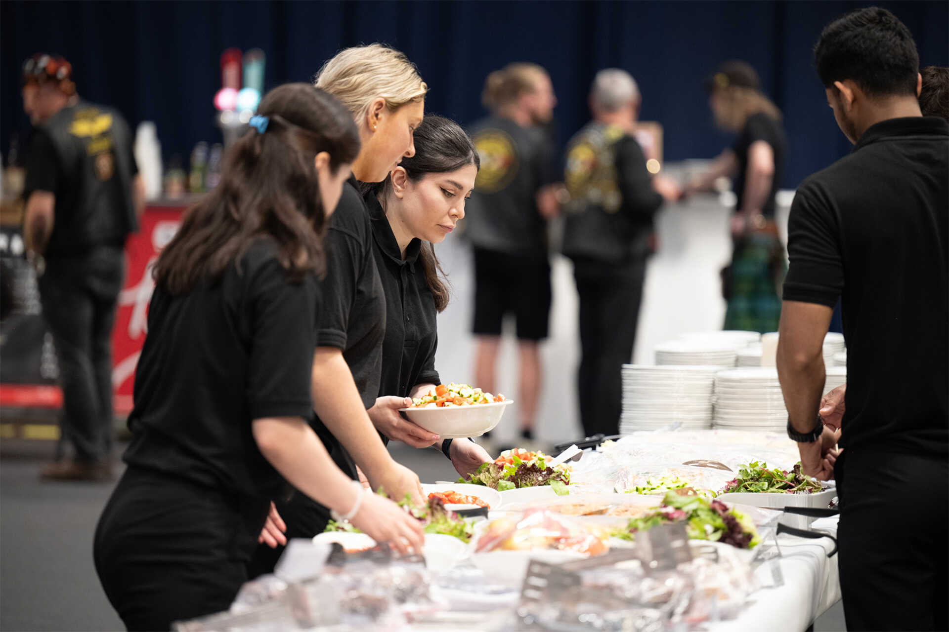 Campus-event guests and staff building salads and plated meals from a buffet setup in a large hall