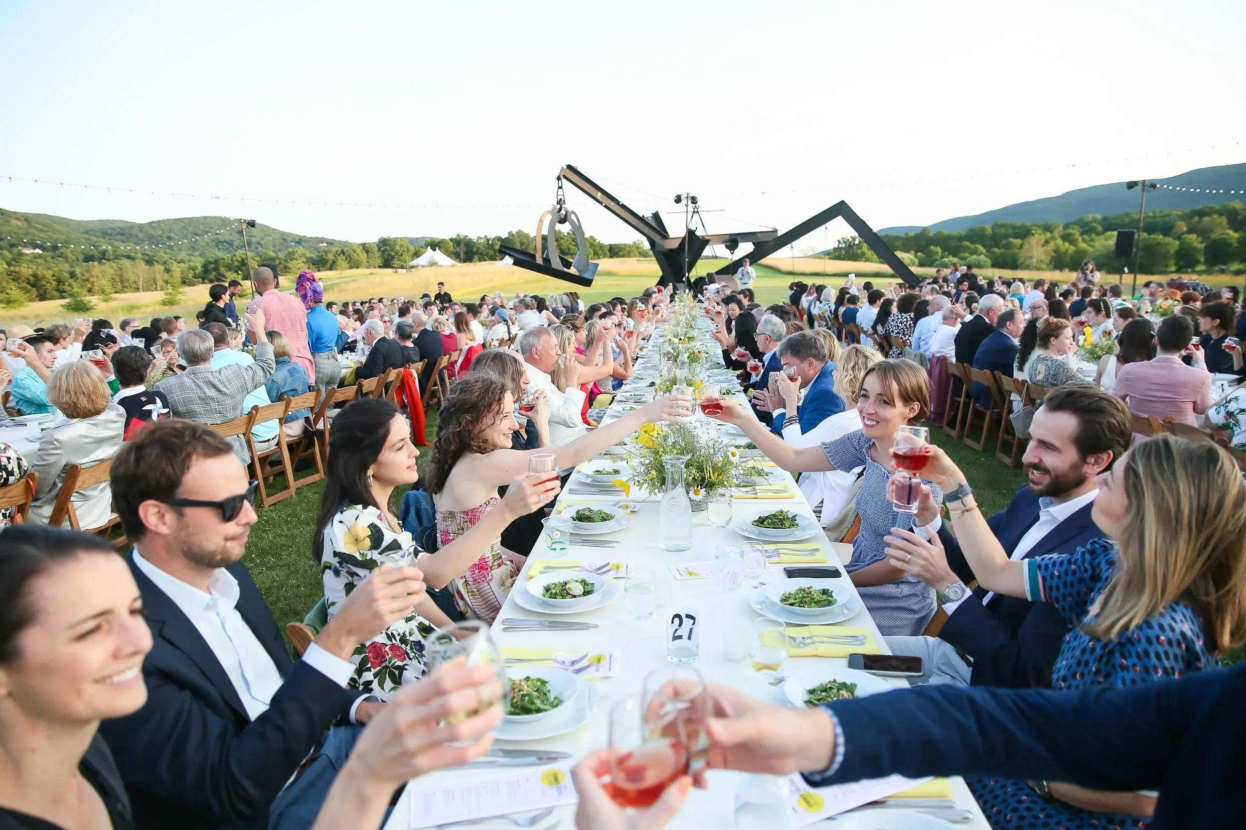 Guests seated along a long outdoor dinner table beneath trees during a garden celebration